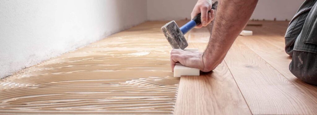 Ein Mann hämmert im Bereich „Über Uns“ mit einem Hammer auf einen Holzboden. Firmenadresse