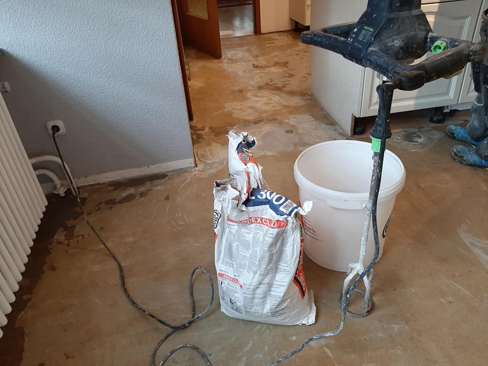 Materialien und Werkzeuge für ein Hausrenovierungsprojekt, darunter Bodenverlegungsmaterialien, ein teilweise verbrauchter Beutel mit Spachtelmasse und ein Mixer, liegen auf dem Boden verstreut. Firmenadresse