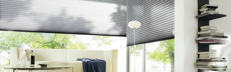 Bild Sonnenschutz in Modernem Wohnzimmer mit großen, mit grauen Rollos behangenen Fenstern, einem cremefarbenen Sofa, einem Couchtisch aus Glas und Regalen voller Bücher mit verschiedenen Produkten. Firmenadresse