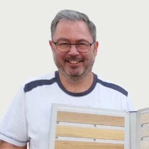 Malermeister Armin Lauer mit Brille und weißem Hemd mit grauen Flicken hält ein Schaubuch mit verschiedenen Holzmustern hoch. Er lächelt in die Kamera und verkörpert damit den freundlichen Geist unserer Rubrik „Über uns“. - Firmenadresse