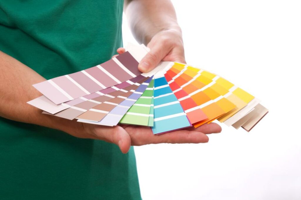 Eine Person hält fächerartig mehrere Farbmuster hoch, auf denen eine Vielfalt an Farben von Rot und Blau bis hin zu Grün und Gelb zu sehen ist. - Firmenadresse