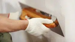 Putz: Eine Person mit weißen Handschuhen glättet mit einer Kelle den Putz einer Wand und schafft so eine ebene Oberfläche, die für einen frischen Anstrich mit Farben-Farbe bereit ist. - Firmenadresse